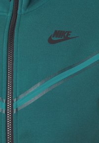 Teal zip-up jacka med svart Nike-logotyp och diagonala reflexrandar över bröstet.