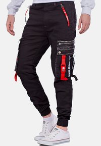 Schwarze Cargo-Hosen mit mehreren Reißverschluss-Taschen, roten Akzenten und Nieten-Details; schmaler Schnitt mit elastischen Bündchen und verstellbaren Riemen.