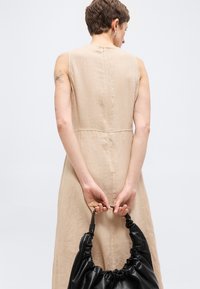 Γυναίκα με κοντά μαλλιά σε μπεζ αμάνικο φόρεμα κρατάει μια μαύρη τσάντα με πιέτες πίσω από την πλάτη, εμφανίζεται από πίσω με φόντο απλό.