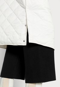 Abrigo acolchado blanco con cierre a presión en el dobladillo, combinado con un short negro texturizado. El diseño enfatiza patrones geométricos y contrastes.