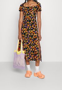 Vestito nero a fiori con maniche a palloncino corte, fiori arancioni e viola, spacco laterale. Tiene in mano una borsa in rete lavanda con dettagli gialli e indossa zoccoli arancioni.