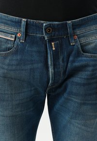 Jeans de mezclilla azul oscuro con un diseño ajustado, que cuentan con herrajes de cobre, costuras visibles y un patrón de decoloración sutil en la tela.