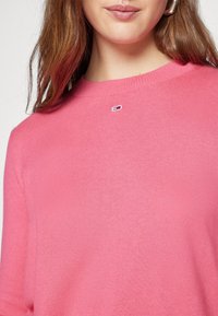 Femme portant un pull col rond rose avec un petit logo rectangulaire au centre, près de la clavicule.
