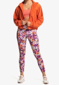 Felpa arancione con zip, crop top rosa, leggings a stampa floreale colorata e sneakers chiare con dettagli texture.