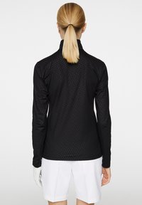 Musta pitkähihainen paita, jossa on teksturoitu kuvio, korkea kaulus ja istuva malli. Yhdistetty valkoisiin shortseihin ja käsineeseen.