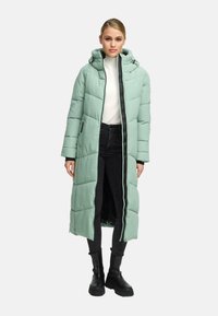 Woman wearing a mint green long puffer coat with hood, white turtleneck, black jeans, and black chunky boots standing against a plain background.