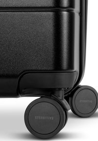Hard-sided black suitcase with a textured surface, featuring four smooth, black wheels, and embossed "ETERNITIV3" branding on wheel caps.