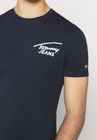 Uomo che indossa una t-shirt Tommy Jeans blu navy con logo bianco sul petto e piccolo logo rettangolare sulla manica.