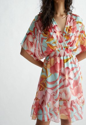 Femme portant une robe bleu clair avec de grands motifs floraux roses et orange, manches courtes, col en V, taille smockée et ourlet à volants.