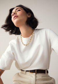 White short-sleeve T-shirt paired with a gold chain necklace and a brown belt, worn with beige high-waisted trousers. Soft texture.