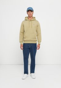 Oliven grønn hettegenser med en frontlomme, snor og logo. Kombinert med blå denimjeans og hvite sneakers. En lyseblå caps fullfører antrekket.
