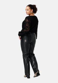 Vrouw met krullend haar in een knot, gekleed in een zwart kanten longsleeve top, zwarte leren broek en hakschoenen met slangenleerprint, zijwaarts gekeerd.