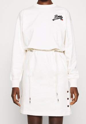 Frau trägt ein weißes, langärmeliges Kleid mit Kordelzug an der Taille, drei seitlichen Knöpfen und dem Schriftzug "Boss Athletic" auf der oberen Brust.