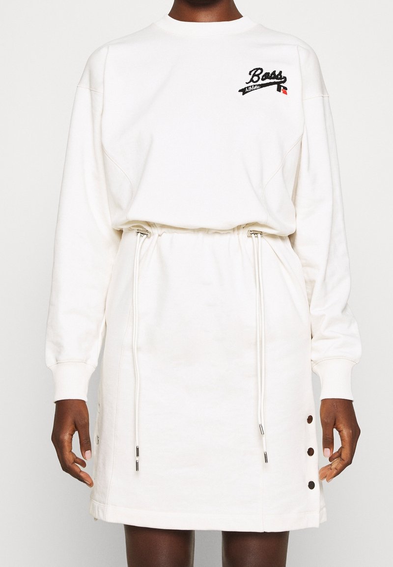 Femme portant une robe blanche à manches longues avec une taille à cordon, trois boutons latéraux et le logo "Boss Athletic" sur la partie supérieure de la poitrine.