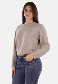 Maglione corto marrone chiaro realizzato in morbido tessuto a maglia, con dettagli di perle sulle spalle e polsini a costine. Abbinato a dei jeans blu.
