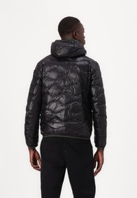 Giacca a vento nera con texture lucida e trapuntata, cappuccio e cuciture verdi a contrasto sui polsini e sull'orlo. Mostra la vista posteriore.