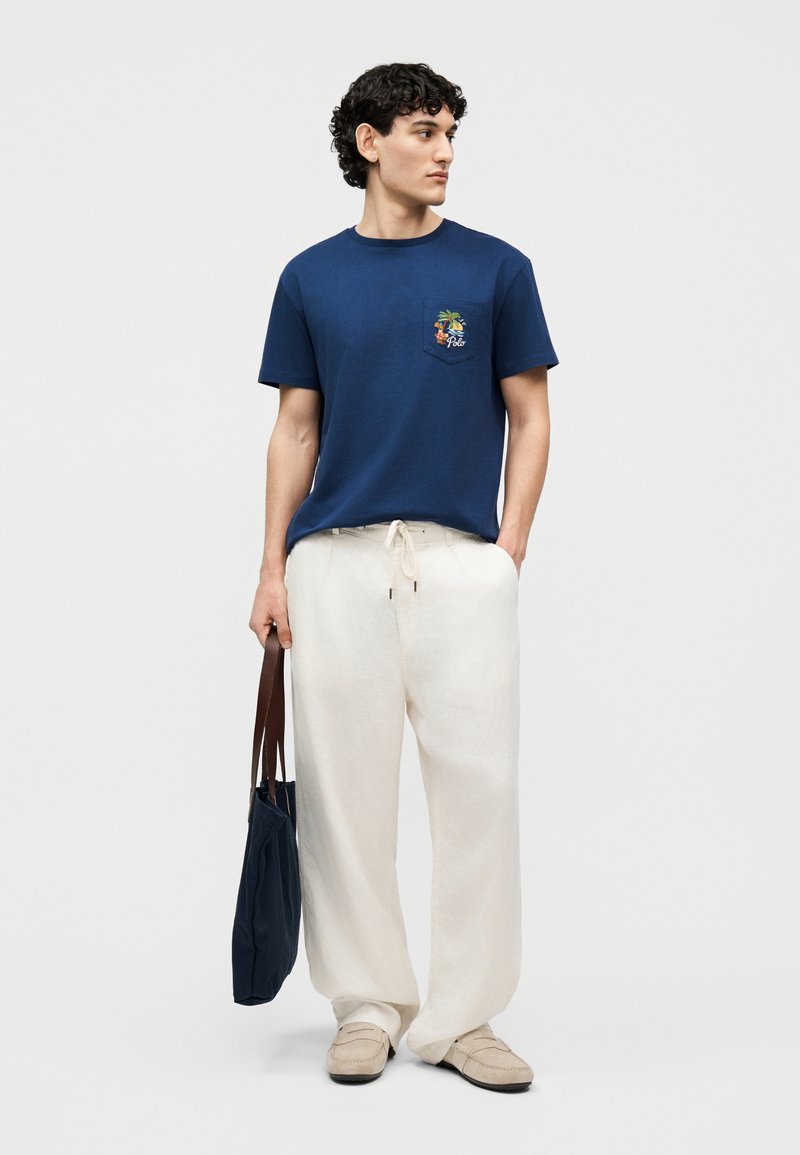 Man in marineblauw T-shirt met tropisch zakontwerp, losse witte broek met trekkoord, beige loafers, en een marineblauwe boodschappentas met bruine bandjes.