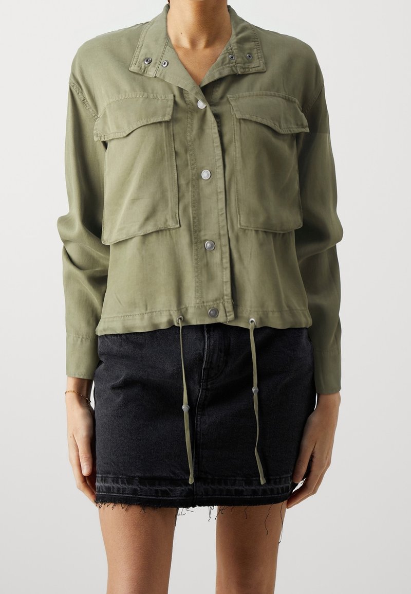 Chaqueta ligera de color verde oliva con mangas largas, dos bolsillos frontales, botones a presión y dobladillo ajustable con cordón, combinada con una falda de mezclilla negra.