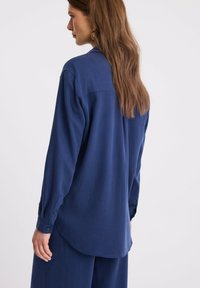 Kobieta z długimi brązowymi włosami, nosząca złote kolczyki oraz luźną, ciemnoniebieską bluzkę z długim rękawem i dopasowane szerokie spodnie, widziana z tyłu.