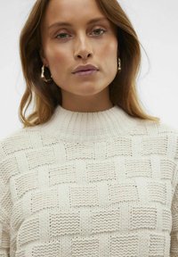 Stickad tröja i off-white med ett strukturerat vävmönster, hög ribbat krage och långa ärmar. Har öppna stygn och grovt garn.