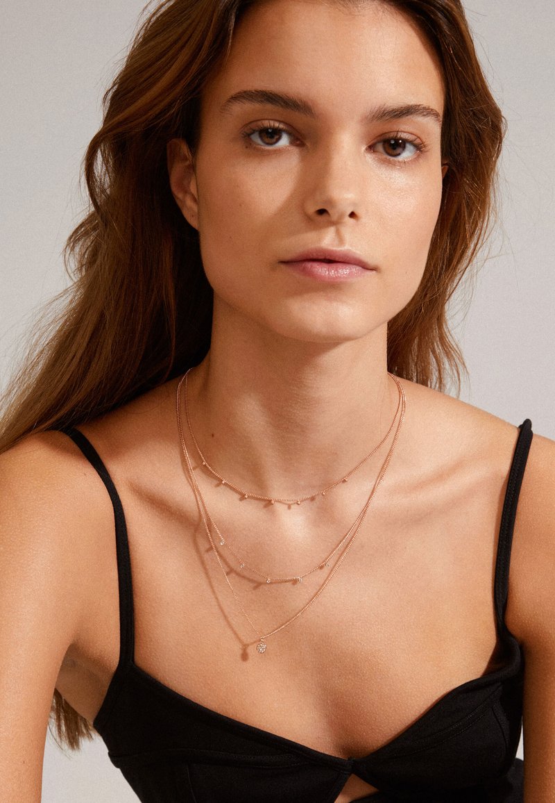 Tres collares en capas en oro rosa. La cadena superior presenta pequeños acentos redondos, mientras que la cadena inferior tiene un pequeño colgante.