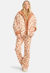 Vrouw die staat in een bijpassende crême en terracotta bloemenpatroon pufferjack en broek met beige schoenen op een witte achtergrond.
