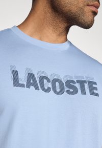 Camiseta de algodón azul claro con un gran logo negro "LACOSTE" en relieve en el frente. Diseño de cuello redondo con mangas cortas.