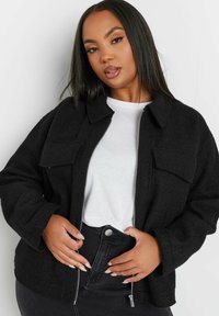 Svart fleecejacka med dragkedja, två bröstfickor, krage, långa ärmar och texturerad yta. Bärs över en vit skjorta och svarta jeans.