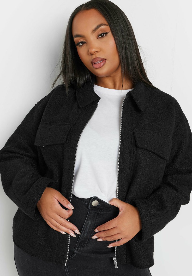 Svart fleecejacka med dragkedja, två bröstfickor, krage, långa ärmar och texturerad yta. Bärs över en vit skjorta och svarta jeans.