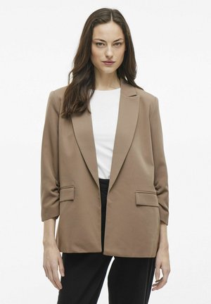 Blazer marrone con chiusura a un bottone, revers a punta, due tasche frontali e maniche a tre quarti, indossato sopra un top bianco.
