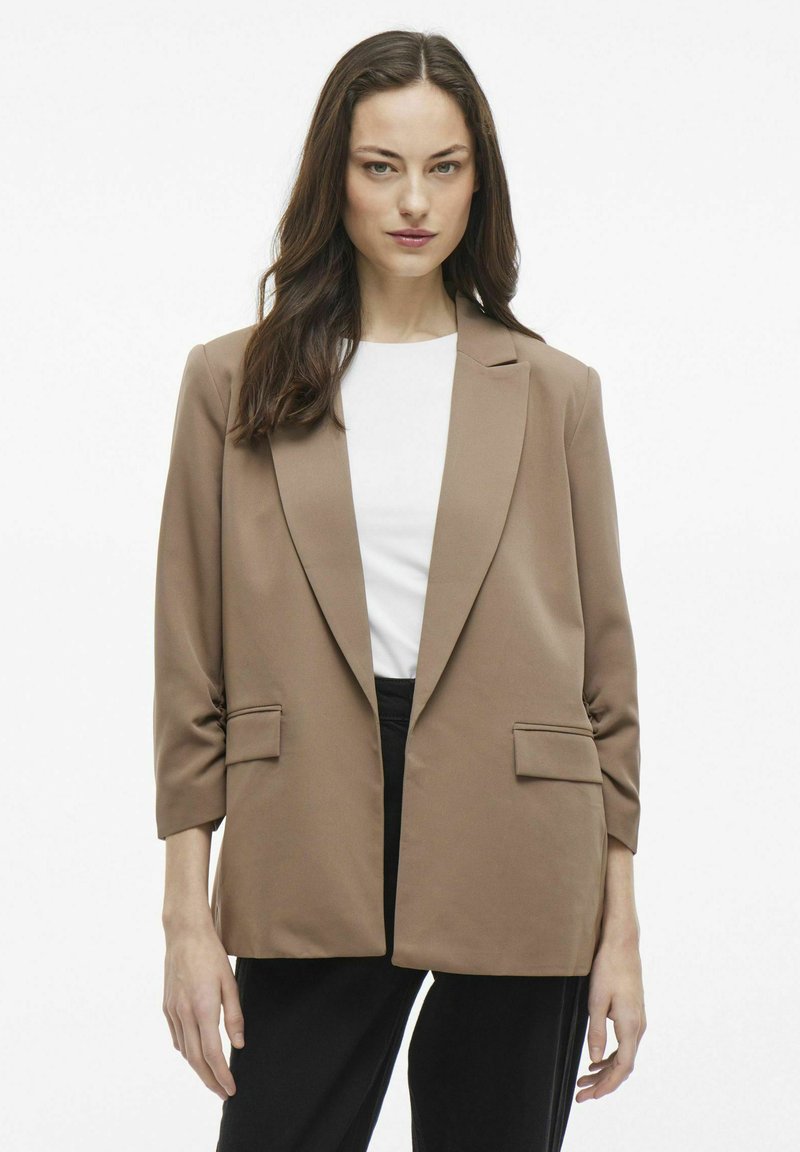 Blazer marrón con cierre de un solo botón, solapas con muesca, dos bolsillos delanteros y mangas de tres cuartos, usado sobre una blusa blanca.