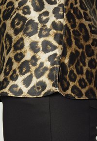 Top s leopardím potiskem s lesklou, saténovou texturou. Dominantními barvami jsou zlatá a černá, s výraznými skvrnami a detaily v podobě bočního rozparku.