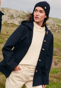 Giacca blu navy con cappuccio, caratterizzata da bottoni in legno e texture trapuntata. Indossata sopra un maglione lavorato a maglia color crema e pantaloni leggeri beige.