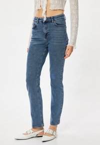 Jeans de mezclilla azules de tiro alto con un corte recto, que presentan un detalle de parche en el muslo izquierdo y un diseño tradicional de cinco bolsillos.