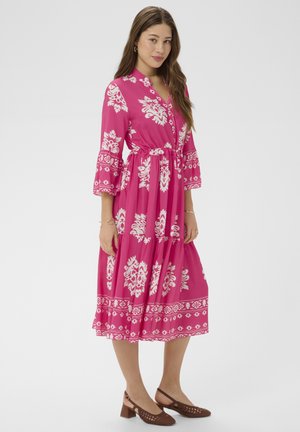 Naine, kes kannab roosat midi-kleit valgete lillemustritega, kolmeveerandvarrukatega ja pruune plokk-kontsadega kootud kingi, seisab ühevärvilise tausta ees.