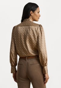 Blusa a maniche lunghe in un tessuto marrone chiaro con un motivo geometrico, dotata di spalle arricciate e colletto alto, abbinata a pantaloni marroni.
