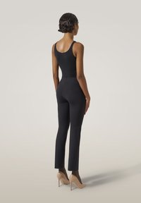 Svart body med tunna band, kombinerad med figurnära svarta byxor. Modellen bär nude högklackade skor med nätstrumpor. Slät textur, minimalistisk design.