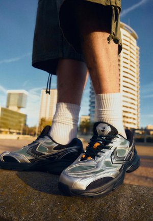 Person trägt silberne und schwarze Sneakers mit weißen Socken und grünen Shorts und steht im Freien mit modernen Gebäuden im Hintergrund.
