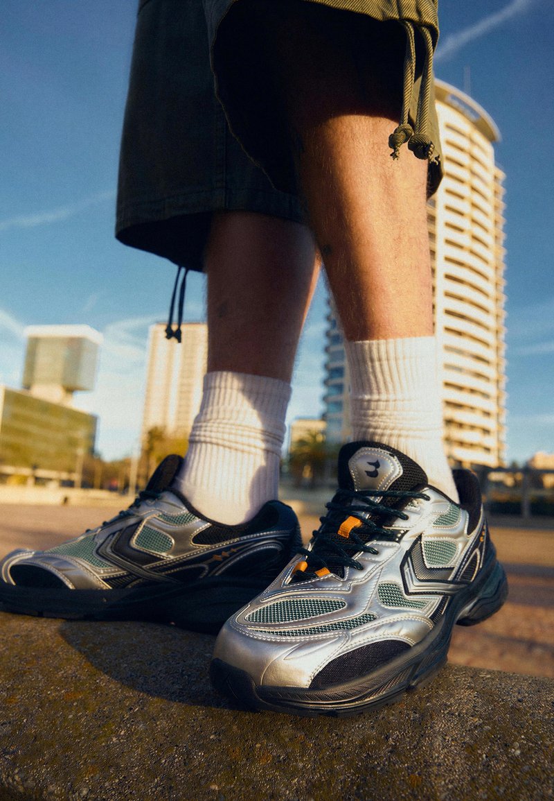 Personne portant des baskets argentées et noires avec des chaussettes blanches et un short vert, debout à l'extérieur avec des bâtiments modernes en arrière-plan.