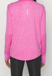 Pitkähihainen urheilupaita kirkkaan vaaleanpunaisena, valmistettu teksturoidusta kankaasta, jossa pyöristetty helma ja valkoinen pystysuora raita takana.