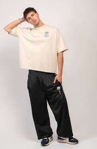 Vīrietis, tērpies bēšā oversized T-kreklā ar tekstu, melnās plašās bikses ar logo un sporta apaviem, stāv ar vienu roku aiz galvas.