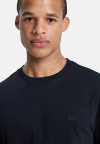 Jeune homme aux cheveux courts portant un t-shirt noir à col rond uni avec un petit logo sur la poitrine gauche, sur fond blanc.
