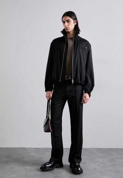 Black zip-up bomber jacket with long sleeves, paired with fitted black pants and chunky black shoes. Model holds a brown handbag.