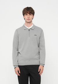 Młody mężczyzna z kręconymi włosami, ubrany w jasnoszary sweter zapinany na zamek z kołnierzem i logo "BOSS" oraz czarne spodnie, stojący na białym tle.