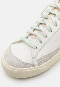 Valkoinen nahkasneakeri, jossa on harmaat mokka-osat, teksturoitu kumipohja, mintunvihreät nauhat ja ompeluyksityiskohdat yläosassa.