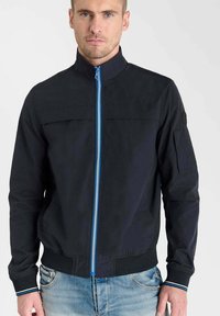 Veste bombardier bleu marine avec un col montant, fermeture éclair à l'avant avec des accents bleus, poches latérales, et poignets et ourlet côtelés. Fabriquée en matériau léger.