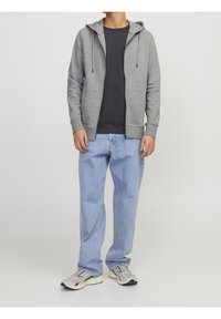 Sudadera con capucha y cremallera en gris con cordones, sobre una camiseta oscura, combinada con vaqueros rectos en azul claro y zapatillas deportivas grises.