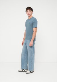 Ανοιχτό μπλε βαμβακερό μπλουζάκι με μικρό τυπωμένο λογότυπο, συνδυασμένο με φαρδιά ξεθωριασμένα τζιν παντελόνια και ανοιχτόχρωμα παπούτσια με σκουρόχρωμες λεπτομέρειες.