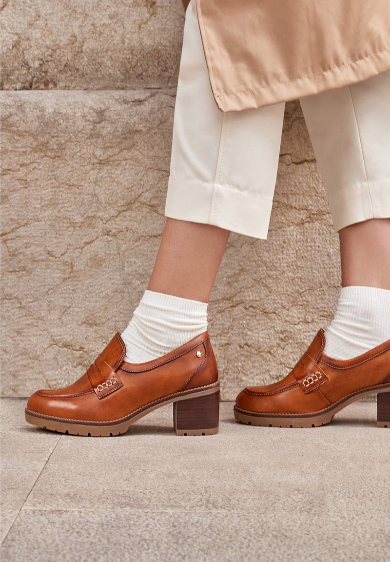Pieds portant des chaussettes blanches et des mocassins en cuir beige à talons blocs, posés sur un sol pavé en pierre devant un mur beige texturé.