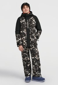 Chaqueta de esquí con capucha en patrón de camuflaje negro y crema, combinada con pantalones de nieve a juego. Botas de nieve azules y un gorro de lana negro completan el atuendo.